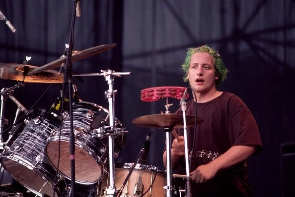 Drummer performing energetically on stage with vibrant hair and casual attire