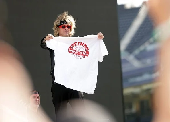 Billie Joe Armstrong holding a large white Green Day t-shirt with a red logo on a stage, wearing a wig, red sunglasses, and a headband