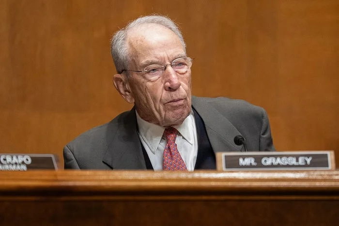 Chuck Grassley is seated, wearing a suit and tie, in a formal setting with his nameplate visible in front