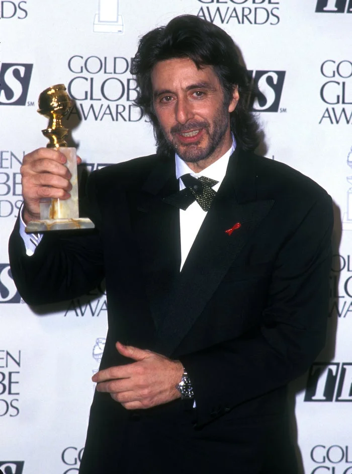 Person in a tuxedo holding a Golden Globe award, standing in front of a backdrop with event logos