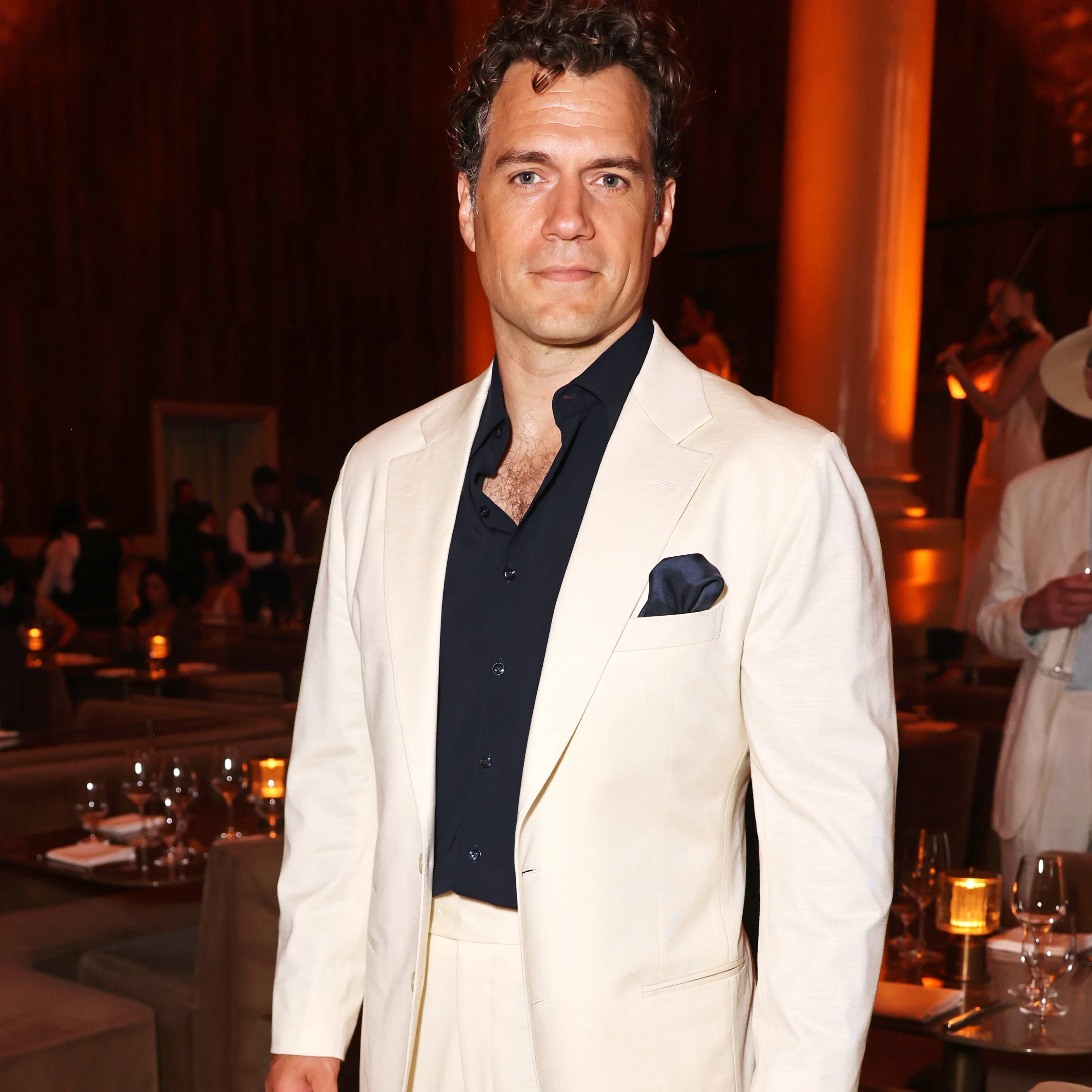A person in a cream suit with a black shirt and pocket square stands in a formal event setting with chandeliers