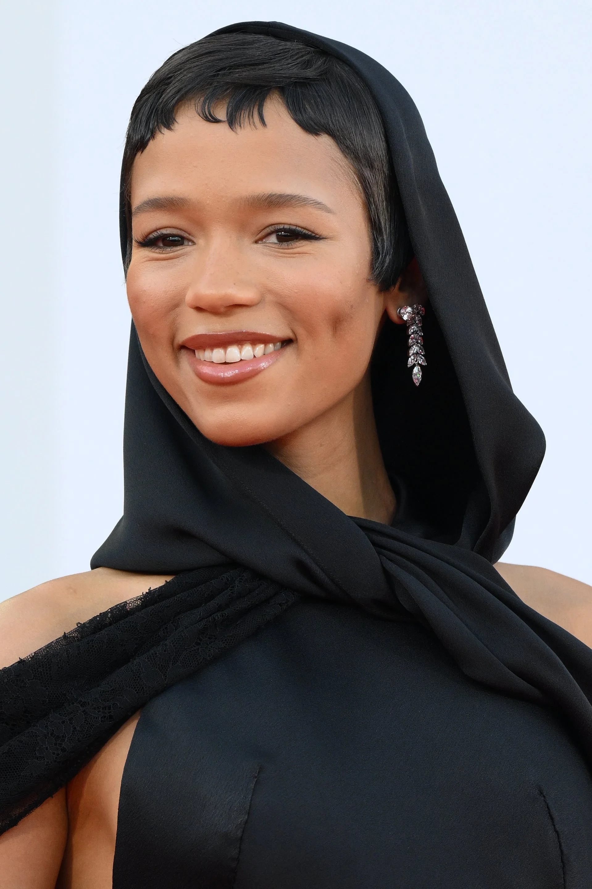 Person in a sleek black gown and headscarf on the red carpet, smiling, with noticeable earrings