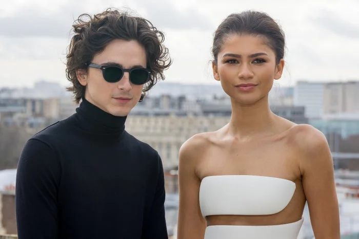 Two individuals pose outdoors. One wears a black turtleneck and sunglasses; the other wears an elegant white outfit with a cutout design