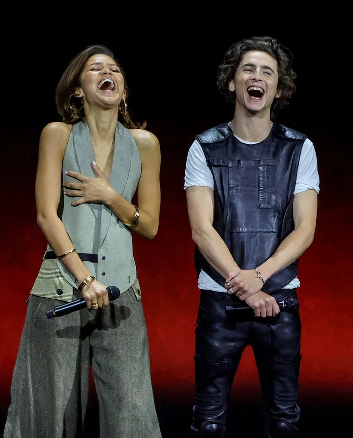 Two people on stage, both laughing. One wears a stylish sleeveless top with wide-legged pants, the other a leather vest over a white T-shirt with leather pants