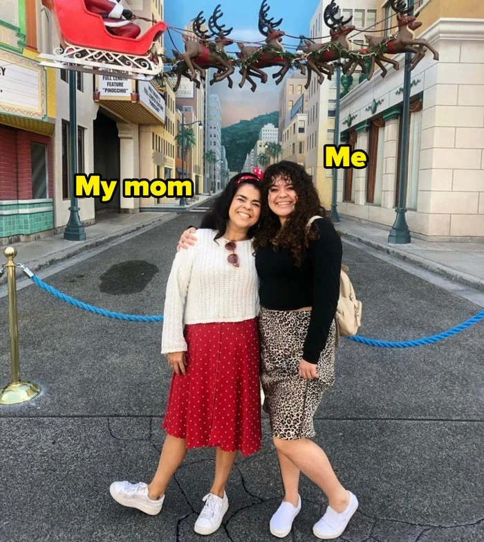 Two people pose happily on a city street backdrop with a Santa sleigh and reindeer overhead. One wears a red skirt, the other a leopard print skirt