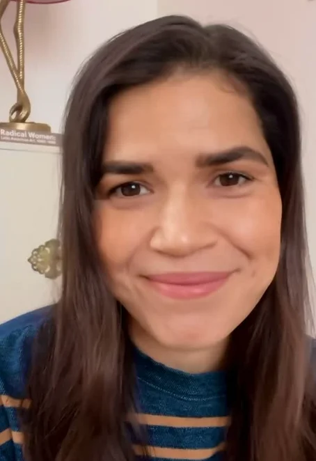 Person smiling, with long hair, wearing a striped sweater. Background shows part of a bookshelf and a small lamp