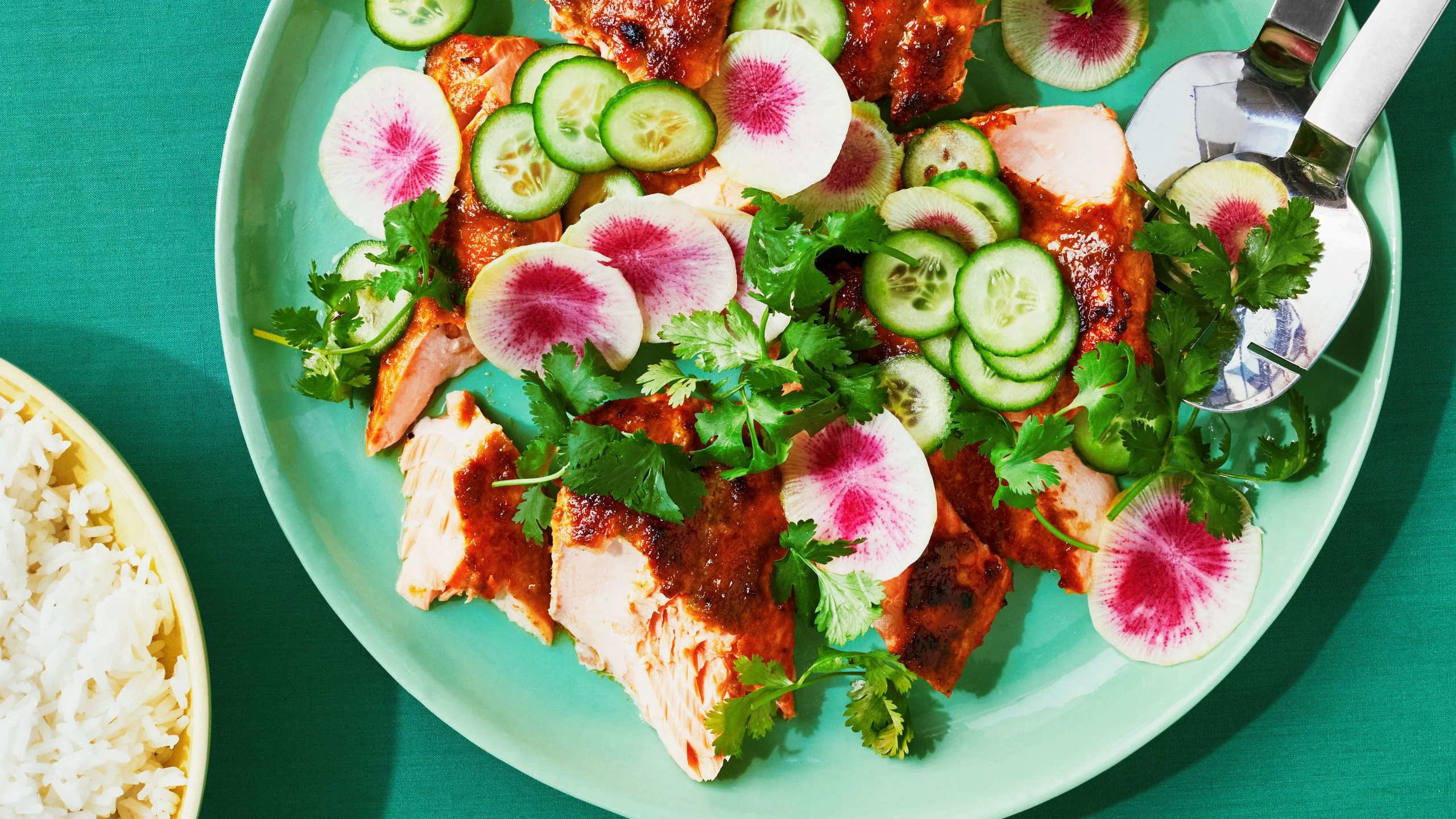 Broken up pieces of broiled salmon covered in a green curry sauce on a platter with sliced cucumbers rainbow radishes...