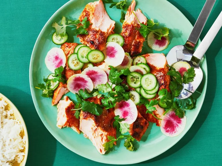 Broken up pieces of broiled salmon covered in a green curry sauce on a platter with sliced cucumbers rainbow radishes...