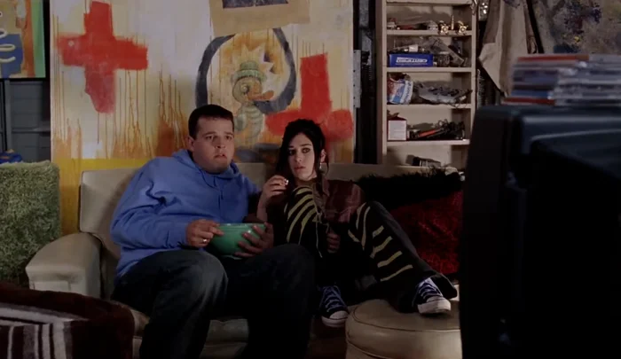 Two people sit on a couch watching TV, one with a bowl of snacks. The room has eclectic decor with art and shelves in the background