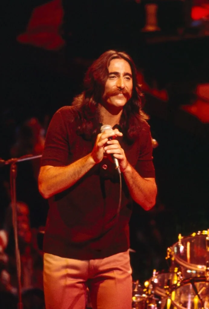 Person on stage holding a microphone, wearing a dark shirt and light pants, with long hair and a mustache, in a performing setting