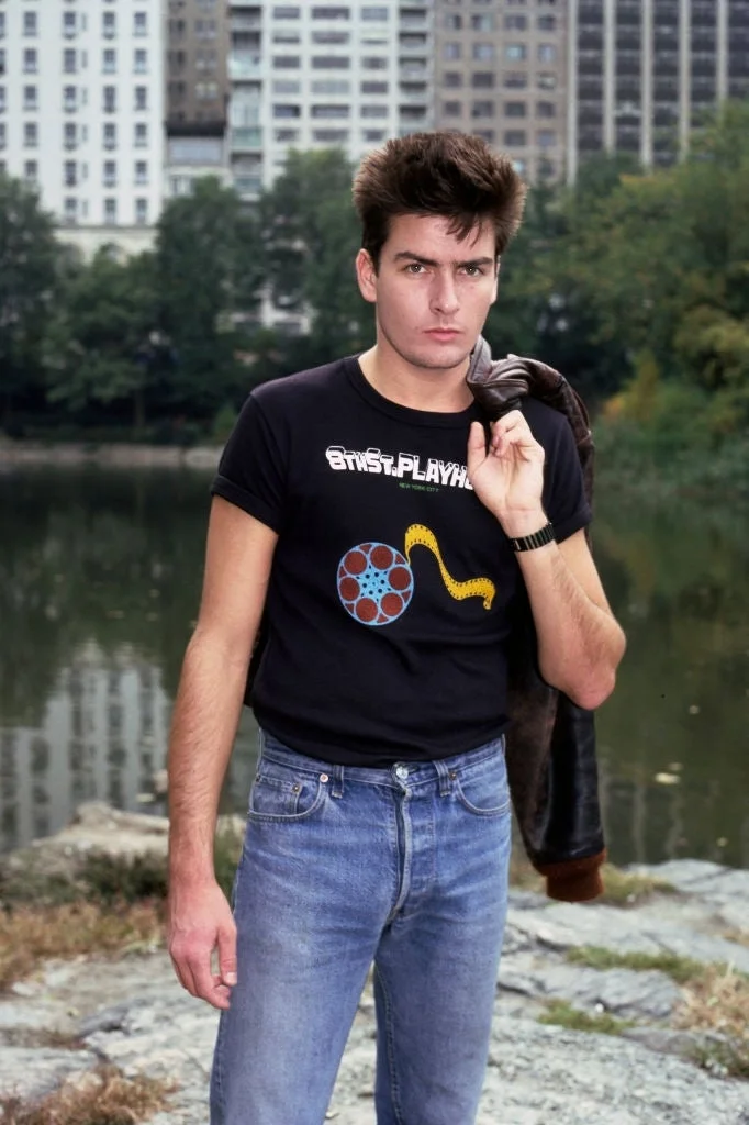 Person outdoors wearing a graphic tee and jeans, holding a jacket over their shoulder with an urban park background