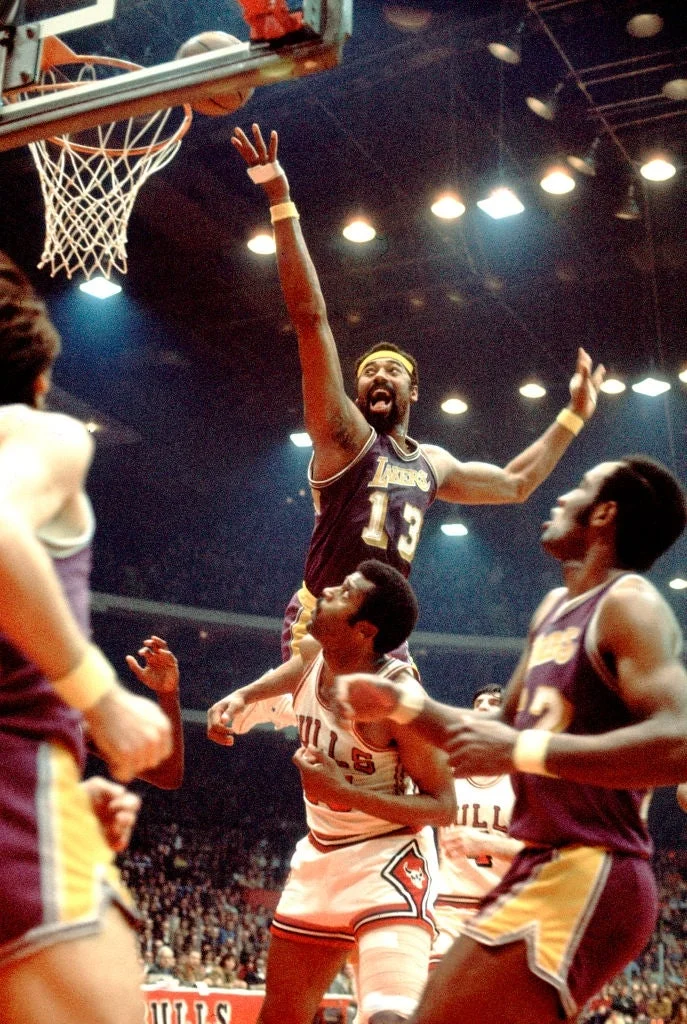 Basketball player in Lakers jersey leaps dramatically over opposing player during a game. Players and audience observe the action