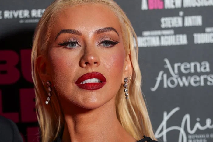 A person with sleek hair, bold makeup, and elegant earrings poses on a red carpet, wearing a stylish outfit