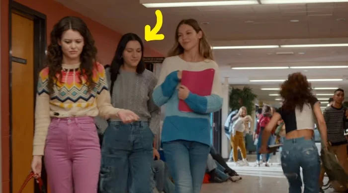 Three people walk down a school hallway, chatting. They wear casual attire: zigzag sweater, gray turtleneck, and color-block sweater. Students in the background