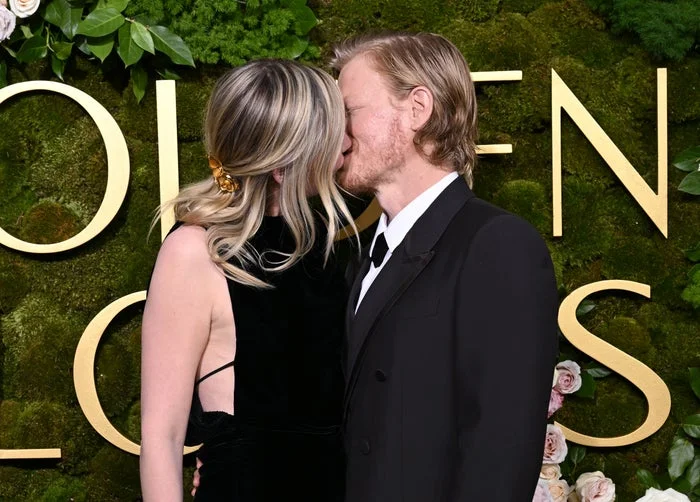 Kirsten Dunst and Jesse Plemons share a kiss on the red carpet. She wears a sleeveless gown, and he wears a suit and tie