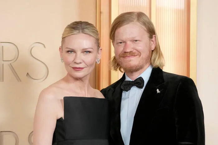 Kirsten Dunst and Jesse Plemons pose elegantly on a formal event carpet. The woman wears a strapless gown, and the man wears a tuxedo with a bowtie