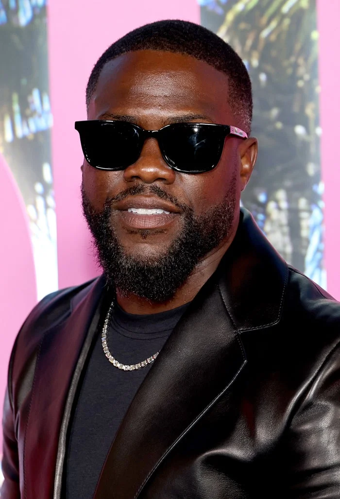 Man in sunglasses wearing a sleek black leather jacket and a necklace, smiling at an event