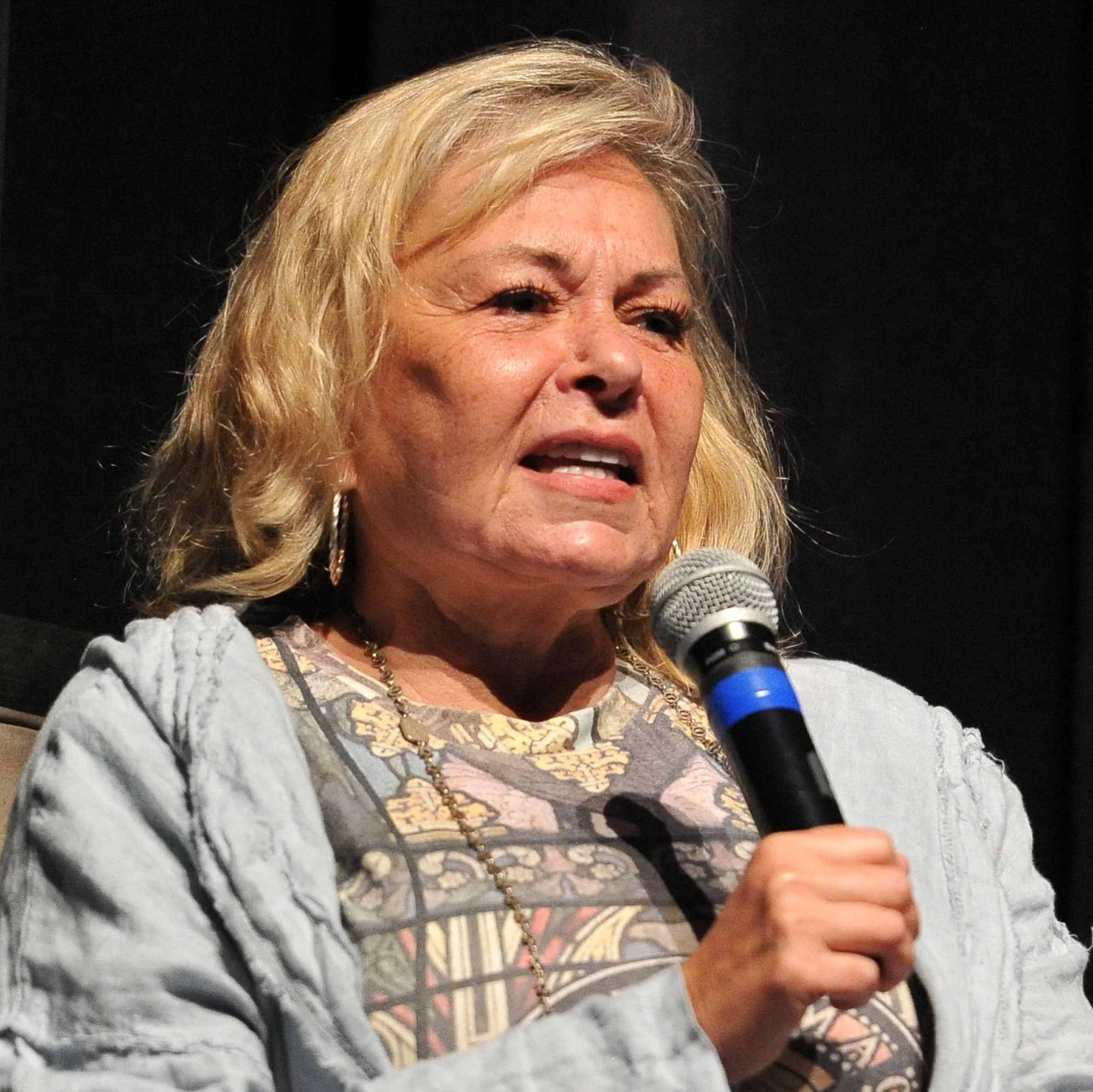 Person speaking into a microphone on stage, wearing a patterned top with a light jacket