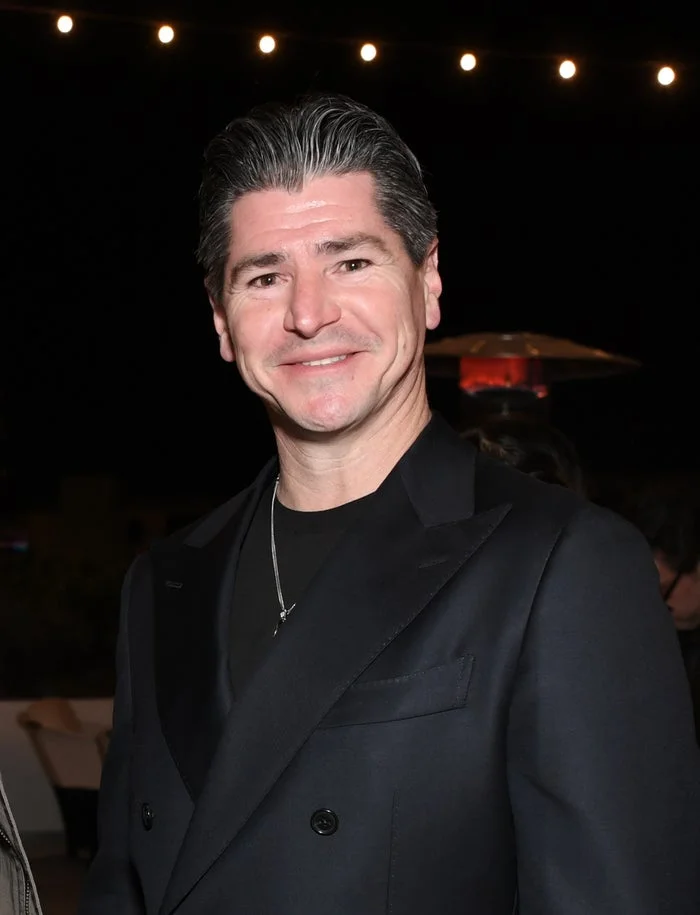 A person in a formal setting, wearing a dark blazer and shirt, smiling at the camera