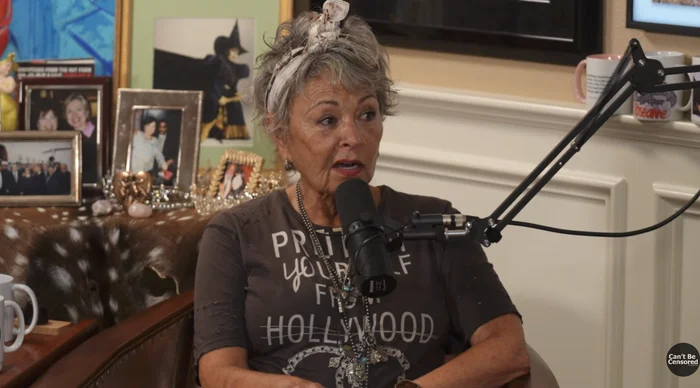 Woman with short hair speaking into a microphone during a podcast. She wears a printed T-shirt and has a headscarf. Background shows framed photos