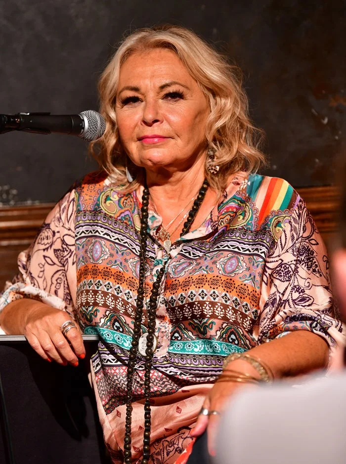 Person with shoulder-length hair in a multicolored, patterned blouse speaks into a microphone at an event