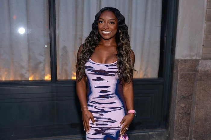 Person in a form-fitting, sleeveless patterned dress, smiling in front of a curtain backdrop at an event