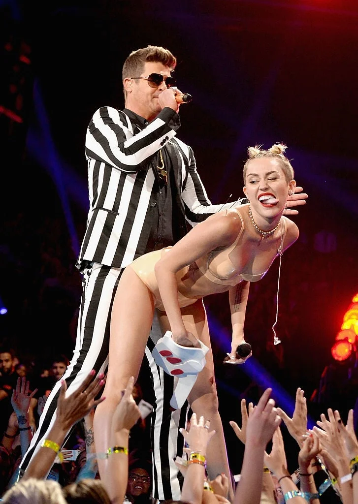 Two performers on stage. One is wearing a striped suit, the other in a minimal outfit with hair buns, posing playfully with visible excitement from audience