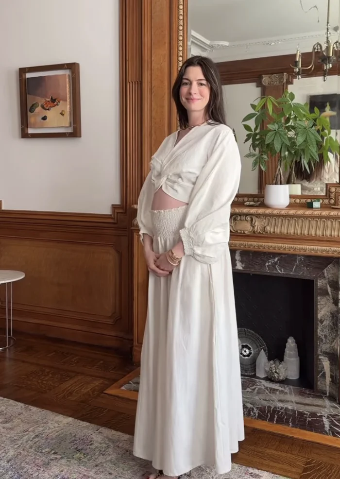 Person in a white gown with a smocked detail, standing in a room with a wooden fireplace and a plant