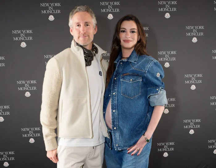 Two people at a New York Moncler event. The woman wears a denim outfit with her pregnant belly visible; the man is in a white jacket and scarf