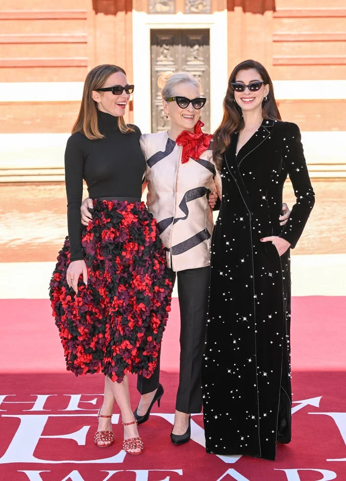 Three women pose together on a red carpet. One wears a floral skirt, another has a floral brooch, and the third dons a starry black gown