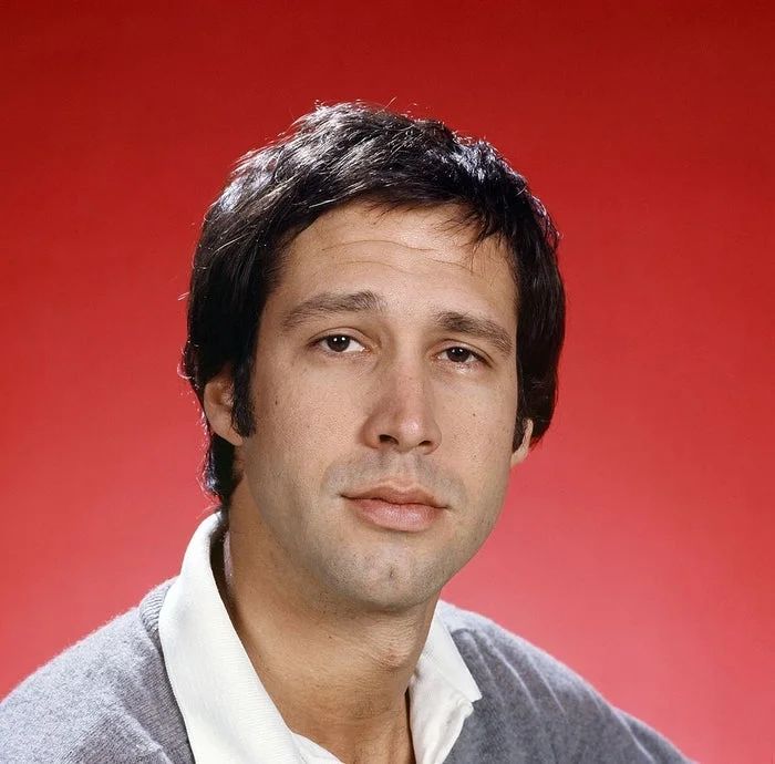 A person with short dark hair wearing a white-collared shirt and a gray sweater against a plain background