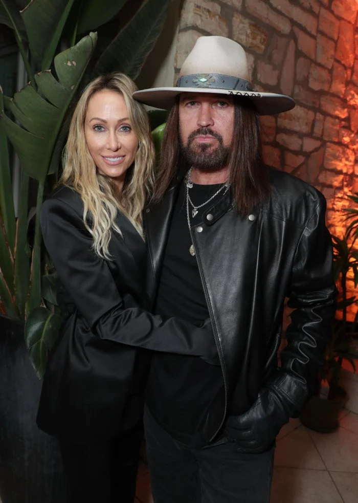 Billy Ray Cyrus and Tish Cyrus pose together against a brick wall with plants in the background. Billy Ray wears a cowboy hat and a leather jacket. Tish wears a black jacket