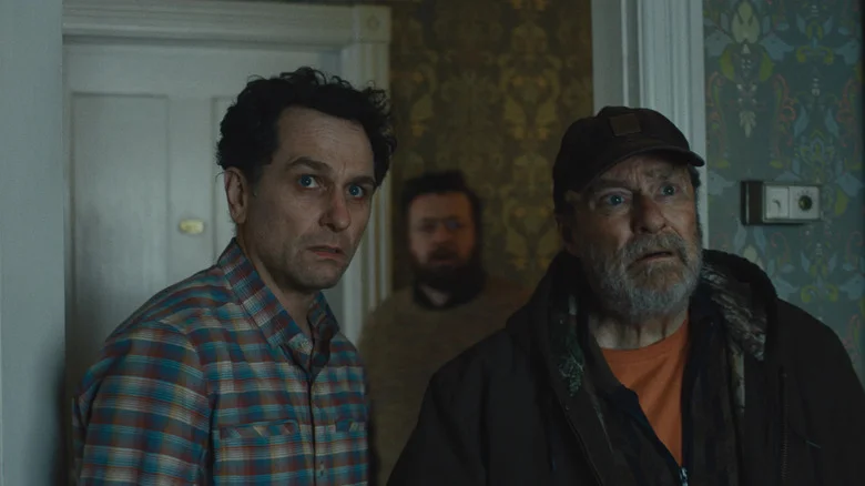 Matthew Rhys as Tom Loftis and Stephen Root as Wyck Crawford in Widow's Bay, standing next to each other with apprehension in a house, with Neil Casey as Kurt standing behind them