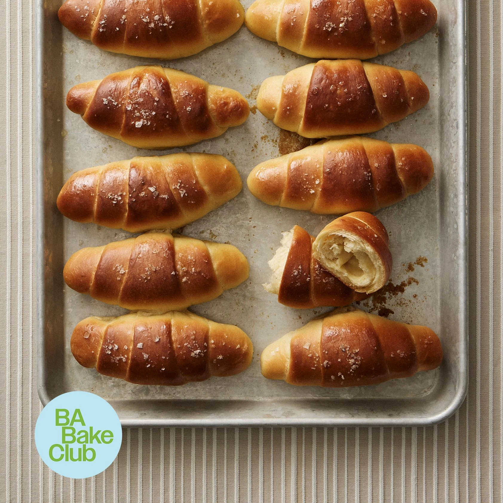 Salt Bread rolls line a metal baking sheet in two columns with coarse salt on top and one roll cut open.