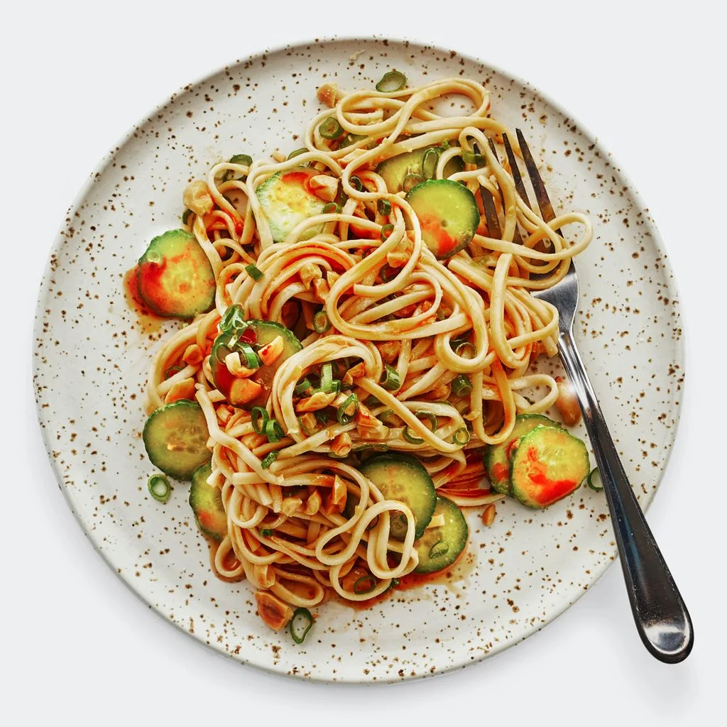 A plate of udon noodles coated in peanut butter sauce and tossed with rounds of cucumber and chopped peanuts drizzled...