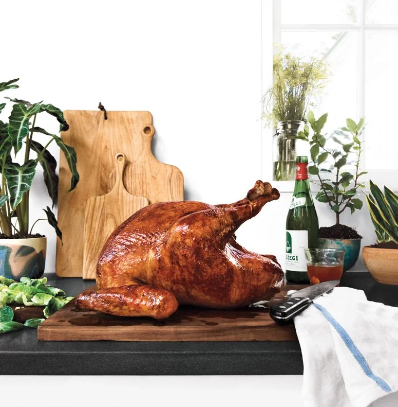 A golden-brown roast turkey on a carving board surrounded by a bottle of wine, brussels spout leaves, a towel, and a carving knife.