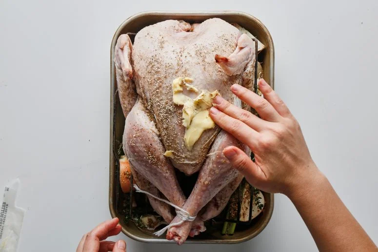 Art for How Long to Cook a Turkey showing a raw, trussed bird in a roasting pan being rubbed with butter.