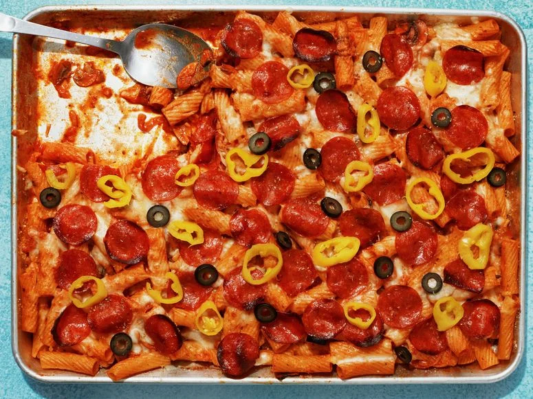 Baked rigatoni with mozzarella pepperoni sliced black olives and sliced pepperoncini on a sheet tray with a serving spoon.