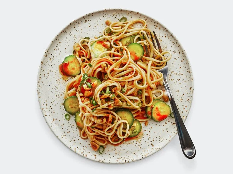 A plate of udon noodles coated in peanut butter sauce and tossed with rounds of cucumber and chopped peanuts drizzled...