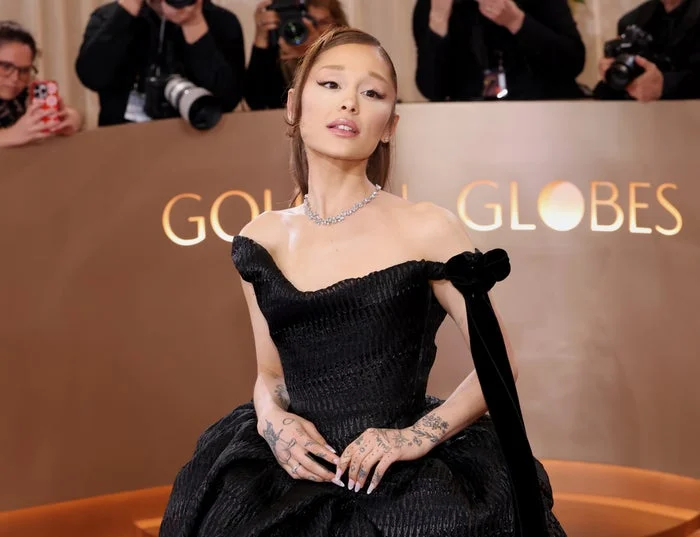 A person poses at the Golden Globes in an elegant off-shoulder, textured black gown with a velvet bow, accompanied by photographers in the background