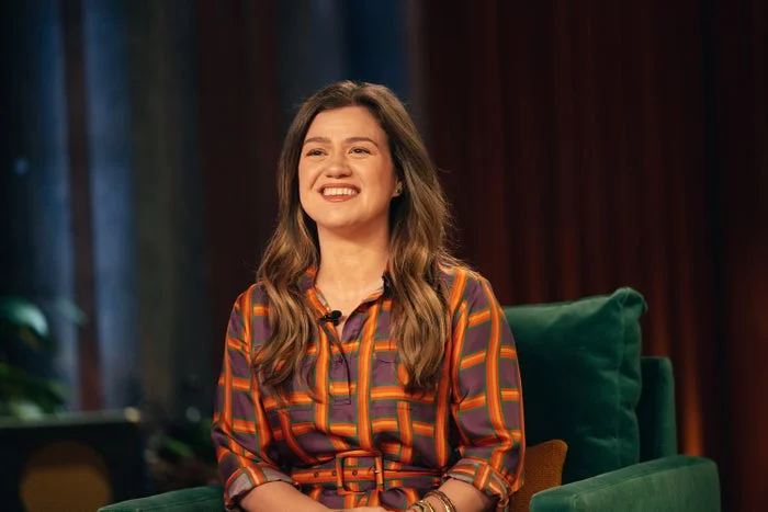 Person sitting on a green chair, smiling, wearing a patterned shirt. Background features indistinct indoor elements