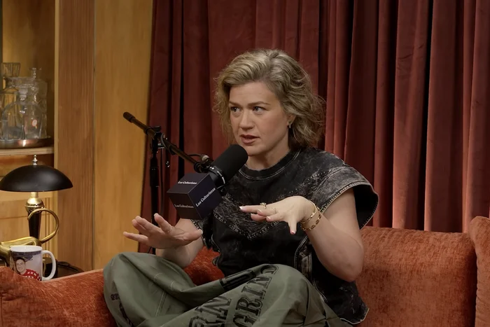 Person sitting on a couch during an interview, gesturing while speaking into a microphone, wearing casual attire