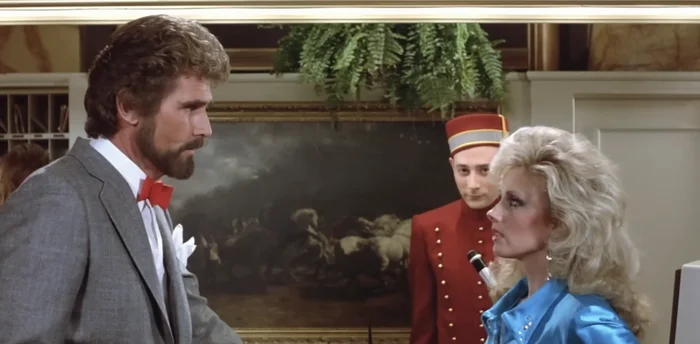 Man in a suit and woman with dramatic hairstyle and bright jacket in a tense conversation at a hotel counter, with a bellboy Pee Wee Herman observing
