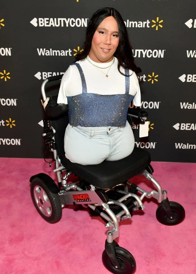 Person in a wheelchair on a pink carpet at an event, wearing a white top with a sequined overtop and light jeans. Sponsors' logos are in the background