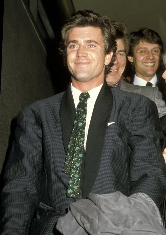 Person in a patterned suit and tie smiles while sitting, with others in the background