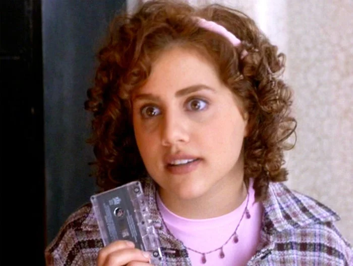 Person with curly hair holds a cassette, dressed in a blazer and necklace, suggesting a retro style