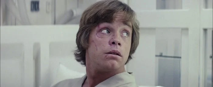 Luke, having short hair looking surprised while seated, wearing a light-colored robe, in a medical or sci-fi setting