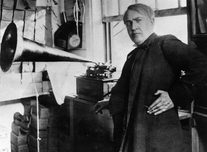 Vintage photo of a man with a phonograph, posed confidently with a cigarette. The setting is a cluttered, industrial space